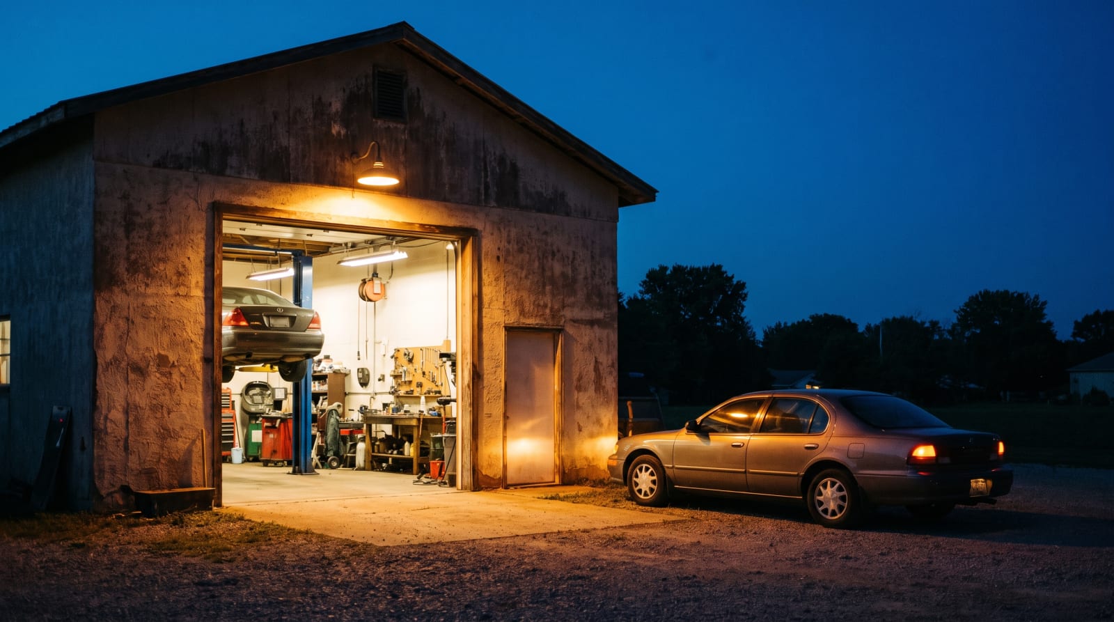 An auto repair shop glowing at dusk with one bay open and a car with hazard lights waiting outside