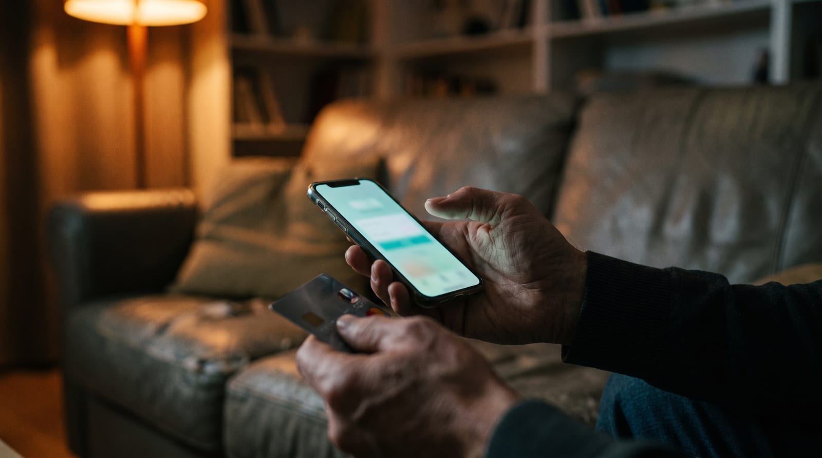 Hands holding a phone at night, thumb hesitating over a checkout screen, credit card in the other hand