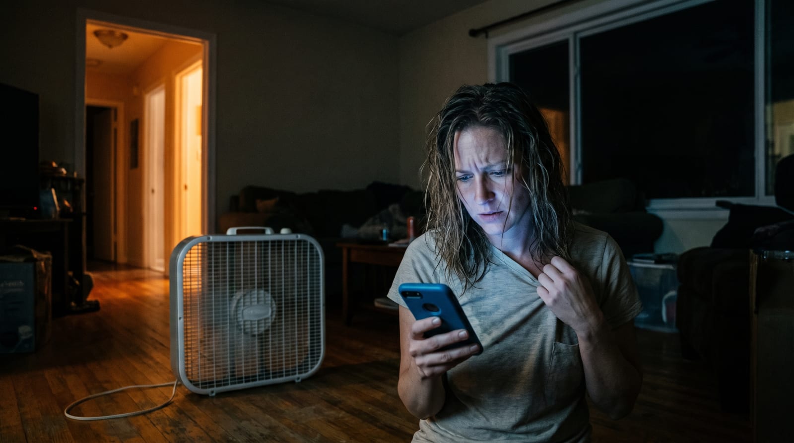 A woman in a dark, hot living room at night urgently searching her phone for emergency AC repair